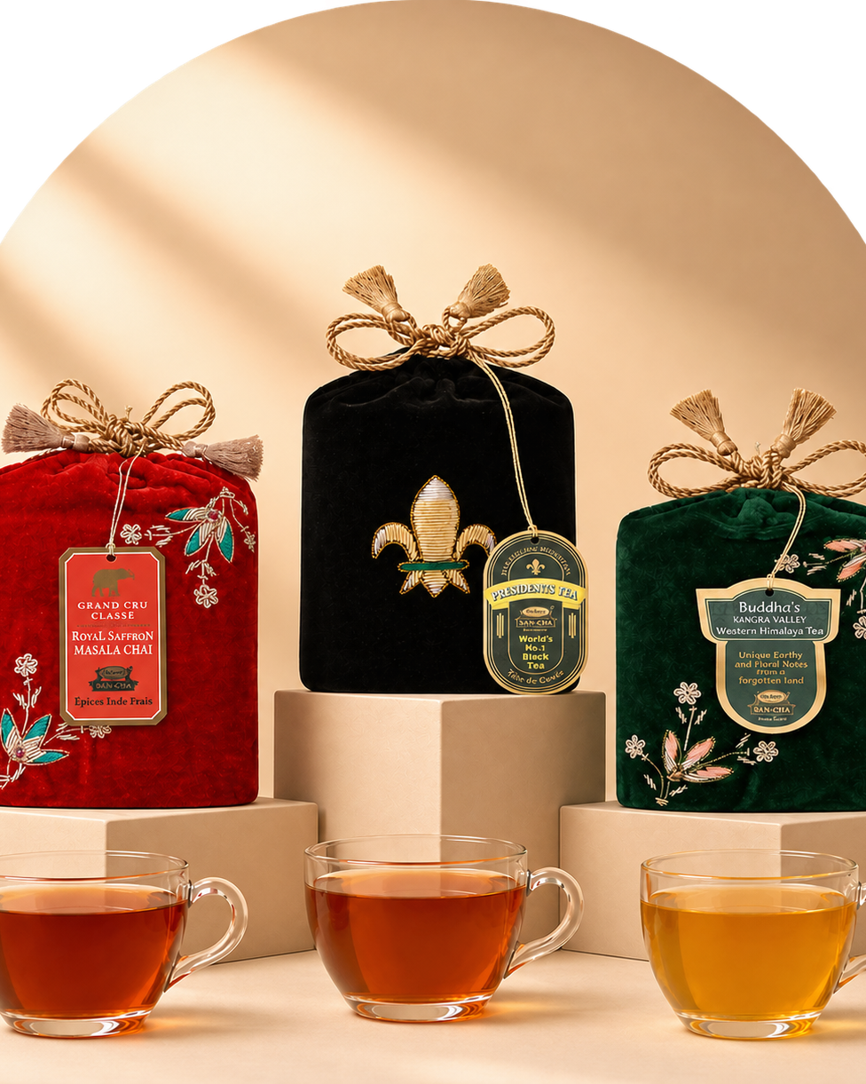 Three packaged tea bags in decorative fabric pouches are displayed on pedestals with three clear glass tea cups in front. The left pouch is red with floral embroidery and a hanging label reading 'The Aromatic Single-Leaf Tea'; the center pouch is black with a gold fleur-de-lis and a round green label; the right pouch is dark green with a cream label reading 'Buddha's Kangra Valley Organic Assam Tea'.