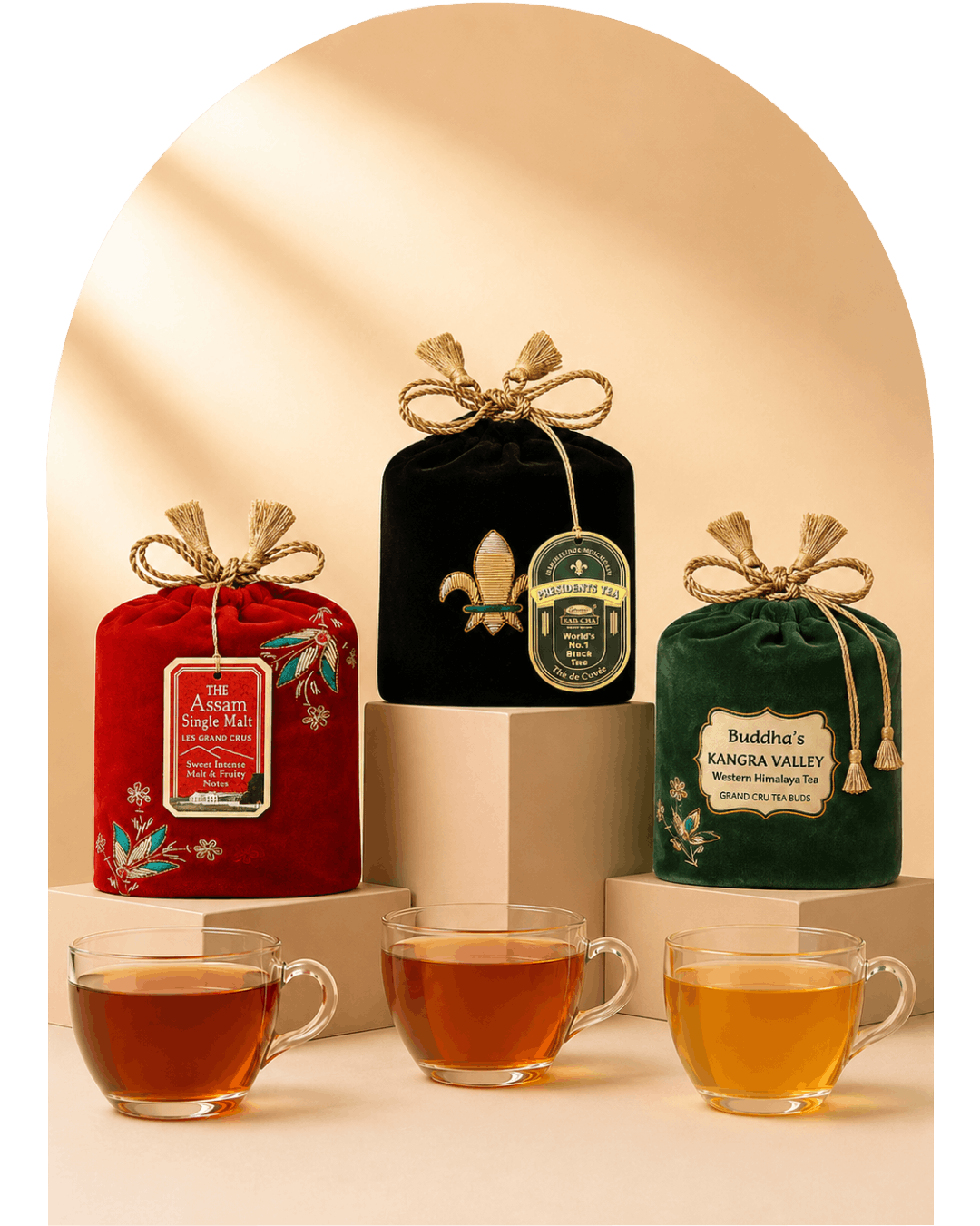 Three packaged tea bags in decorative fabric pouches are displayed on pedestals with three clear glass tea cups in front. The left pouch is red with floral embroidery and a hanging label reading 'The Aromatic Single-Leaf Tea'; the center pouch is black with a gold fleur-de-lis and a round green label; the right pouch is dark green with a cream label reading 'Buddha's Kangra Valley Organic Assam Tea'.