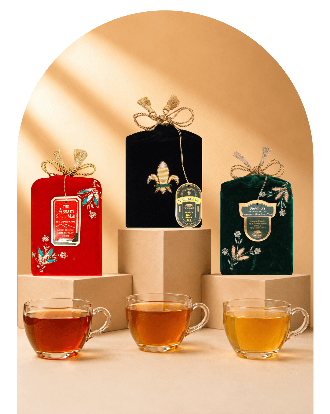 Three packaged tea bags in decorative fabric pouches are displayed on pedestals with three clear glass tea cups in front. The left pouch is red with floral embroidery and a hanging label reading 'The Aromatic Single-Leaf Tea'; the center pouch is black with a gold fleur-de-lis and a round green label; the right pouch is dark green with a cream label reading 'Buddha's Kangra Valley Organic Assam Tea'.
