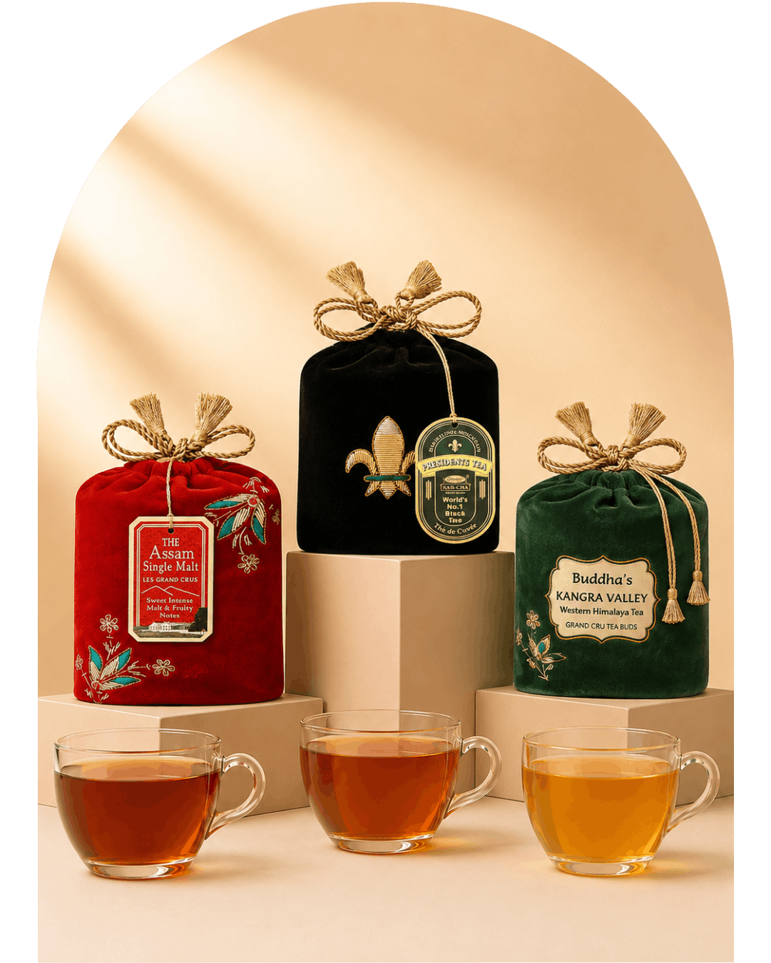 Three packaged tea bags in decorative fabric pouches are displayed on pedestals with three clear glass tea cups in front. The left pouch is red with floral embroidery and a hanging label reading 'The Aromatic Single-Leaf Tea'; the center pouch is black with a gold fleur-de-lis and a round green label; the right pouch is dark green with a cream label reading 'Buddha's Kangra Valley Organic Assam Tea'.